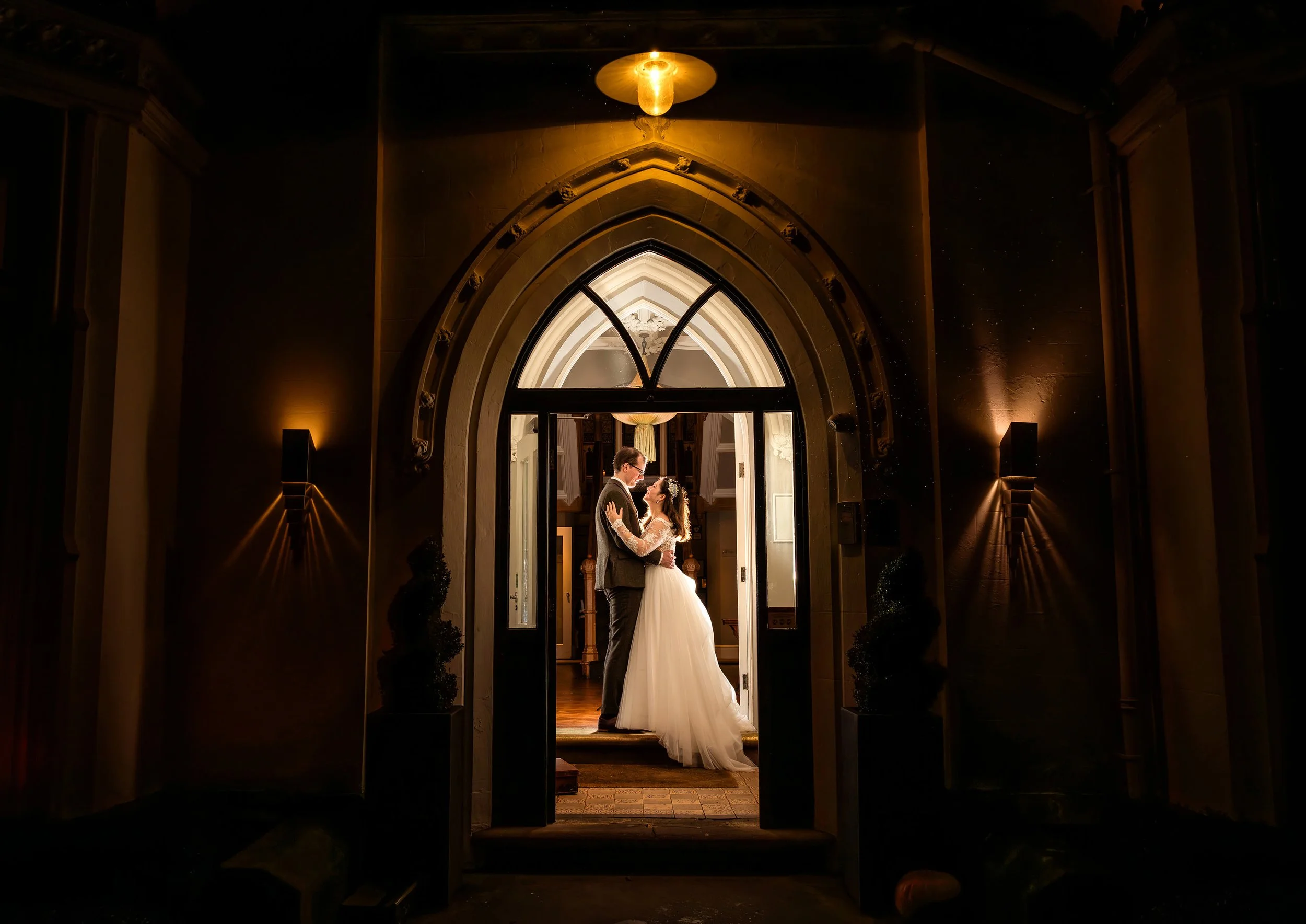A bride and groom are standing close together in a warmly lit, Gothic-style archway at their wedding. The bride is wearing a white gown with lace sleeves and the groom is in a dark suit. They are gazing at each other, creating a romantic scene.