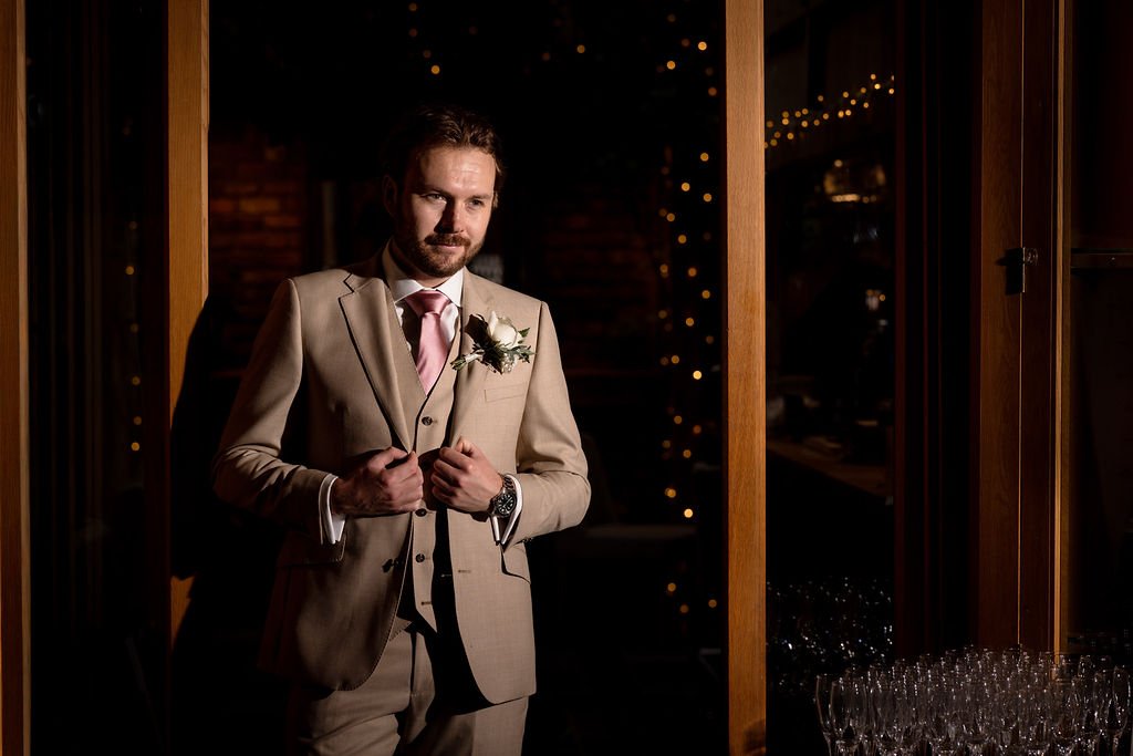 A man dressed in a beige suit and pink tie, standing in a dimly lit room with string lights, holding his lapels, with a boutonnière on his lapel, possibly at a wedding or formal event.