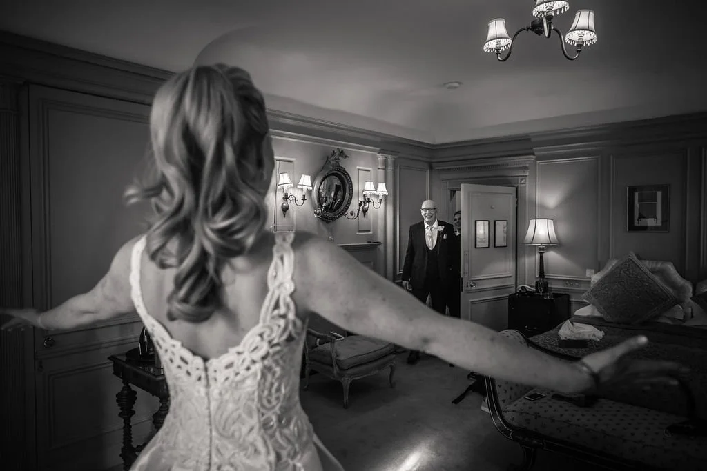 A woman in a wedding dress with outstretched arms standing in a bedroom, facing two men in suits who are smiling and entering the room.