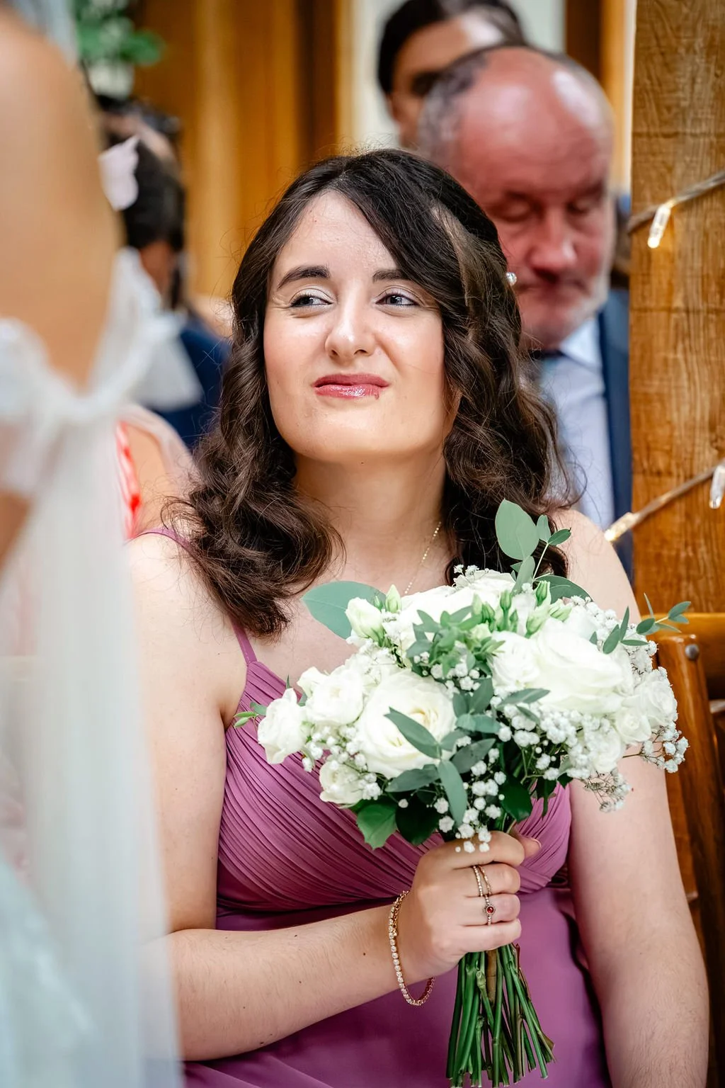 A woman in a purple dress holding a white flower bouquet, smiling slightly at a wedding or celebration event.