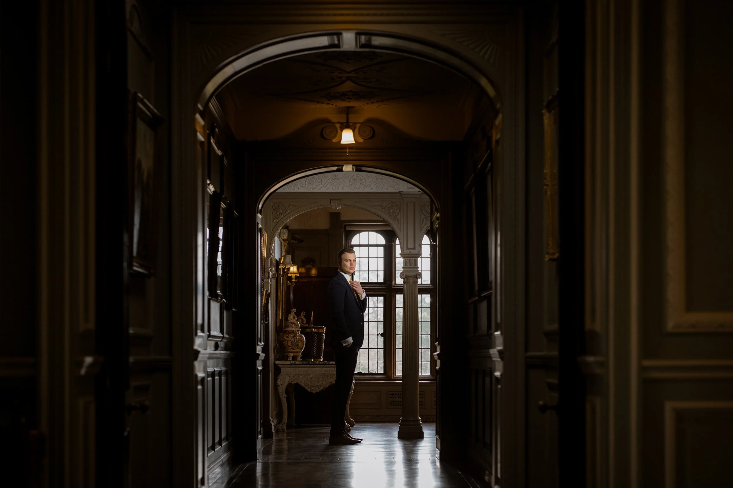 A man in a formal suit standing in a hallway of a historic mansion or hotel, with ornate wooden paneling, decorative columns, and large windows, looking at the camera.