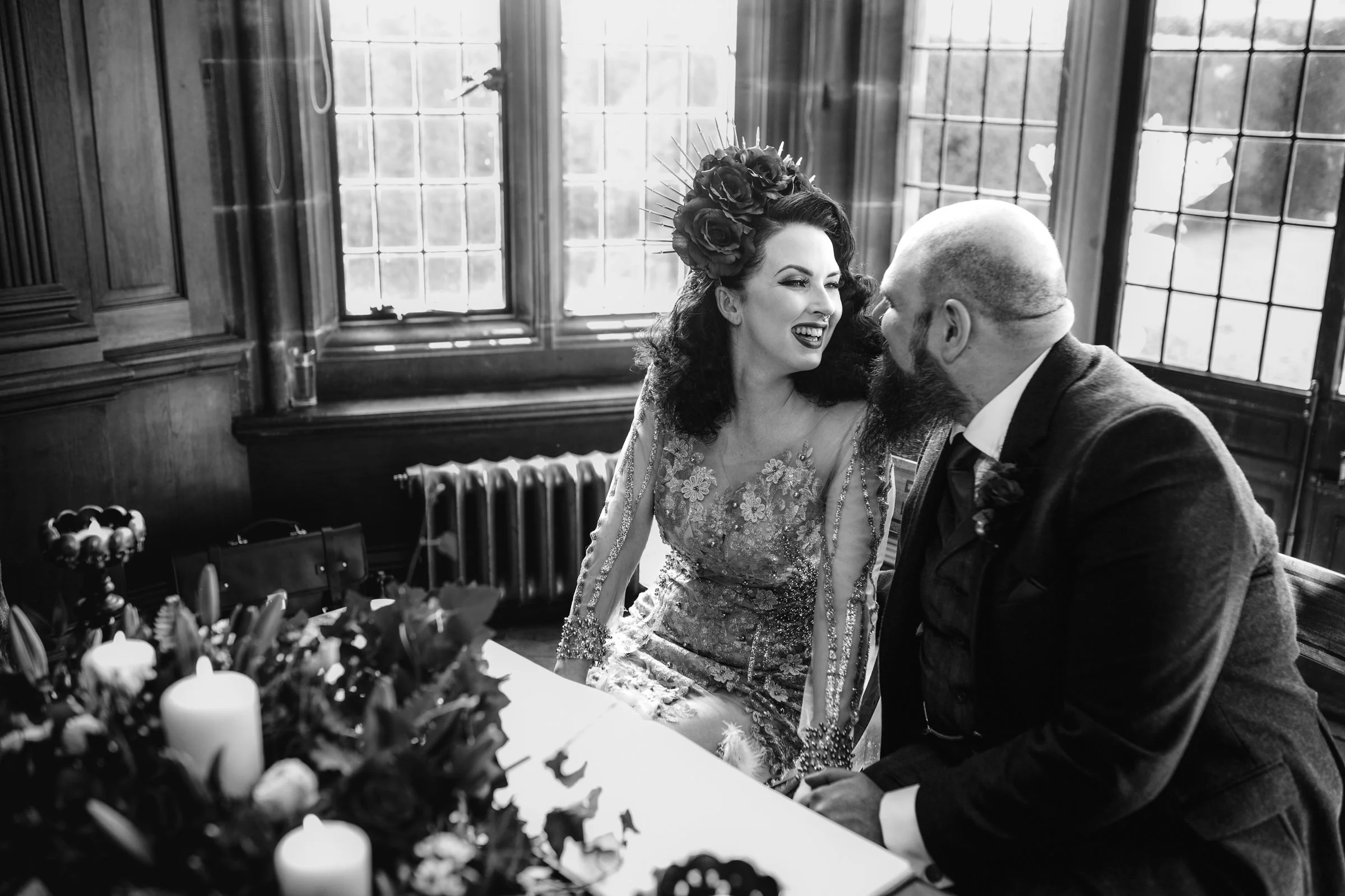 A woman with dark, curly hair, wearing a floral lace dress and a floral headpiece, sitting next to a man with a beard and bald head, dressed in a dark suit, sharing a joyful moment in a vintage-style room with large windows and wooden paneling.