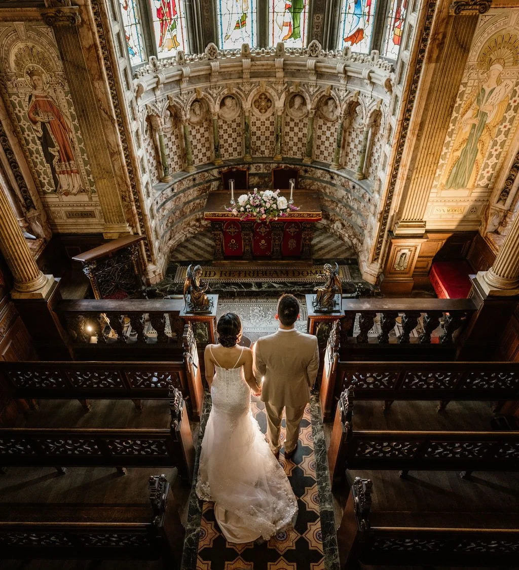 A bride and groom walk hand-in-hand down the aisle of a church decorated for a wedding. The church features ornate stained glass windows, intricate woodwork, and religious statues.