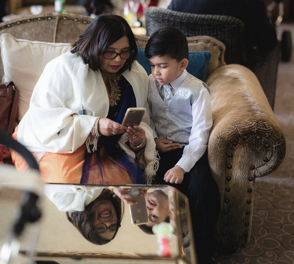 A woman and a young boy are sitting on a beige patterned couch, looking at a smartphone together. The woman wears glasses and a white shawl, while the boy is dressed in a silver vest and white shirt. There are pillows behind them, and a reflective su