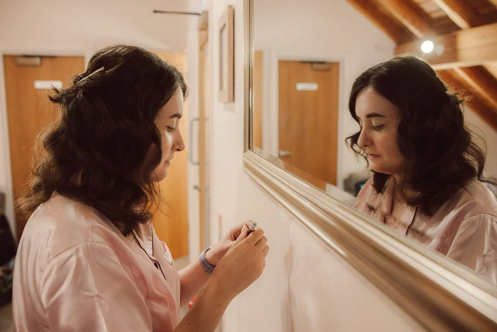 Woman with dark wavy hair and light pink attire writing on a wall, with her reflection visible in a large mirror.