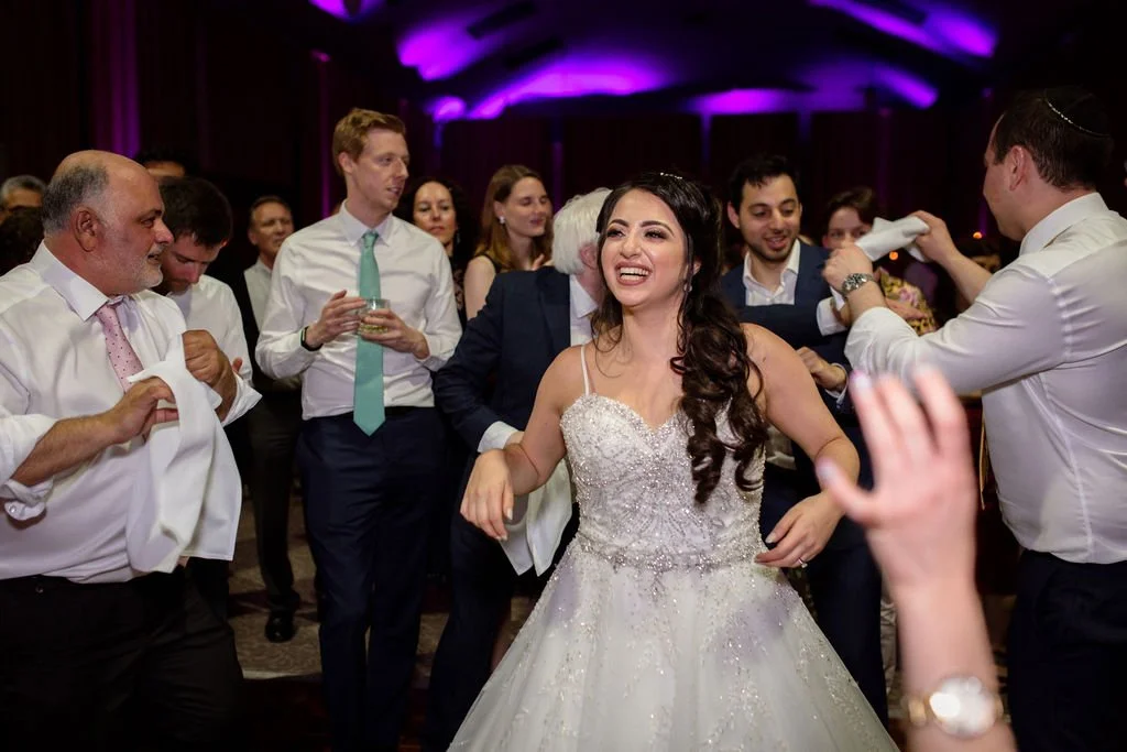 Bride dancing and smiling with wedding guests at reception, purple lighting in background.