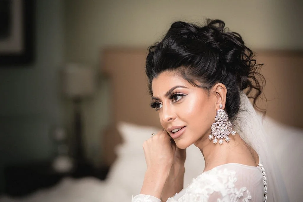 Close-up of a woman with dark, curly hair and large chandelier earrings, wearing a white bridal dress in a softly lit room.
