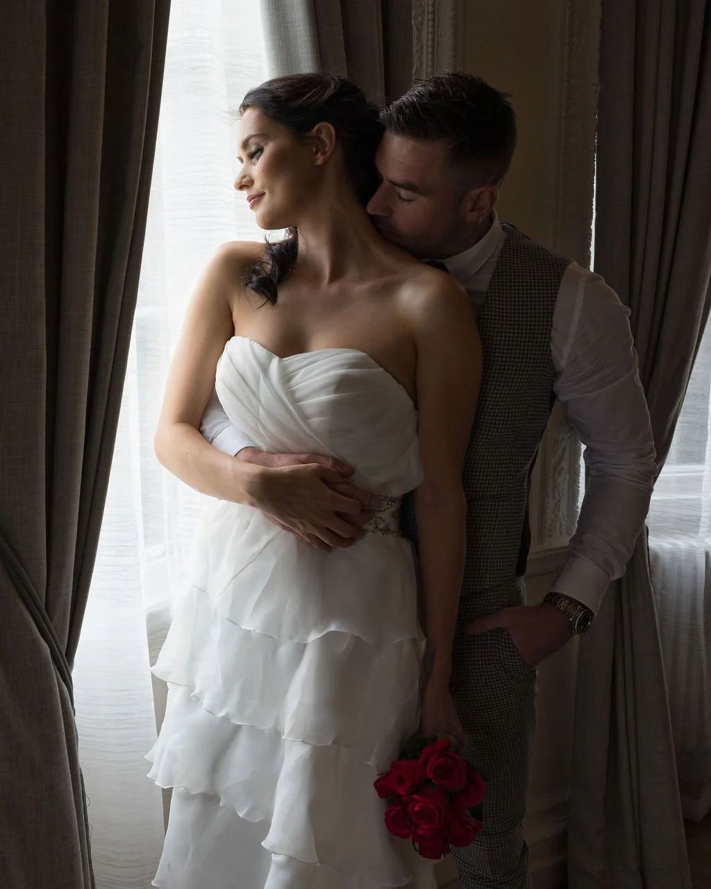 A couple in wedding attire standing near a window, with the groom kissing the bride's neck and the bride holding a bouquet of red roses.