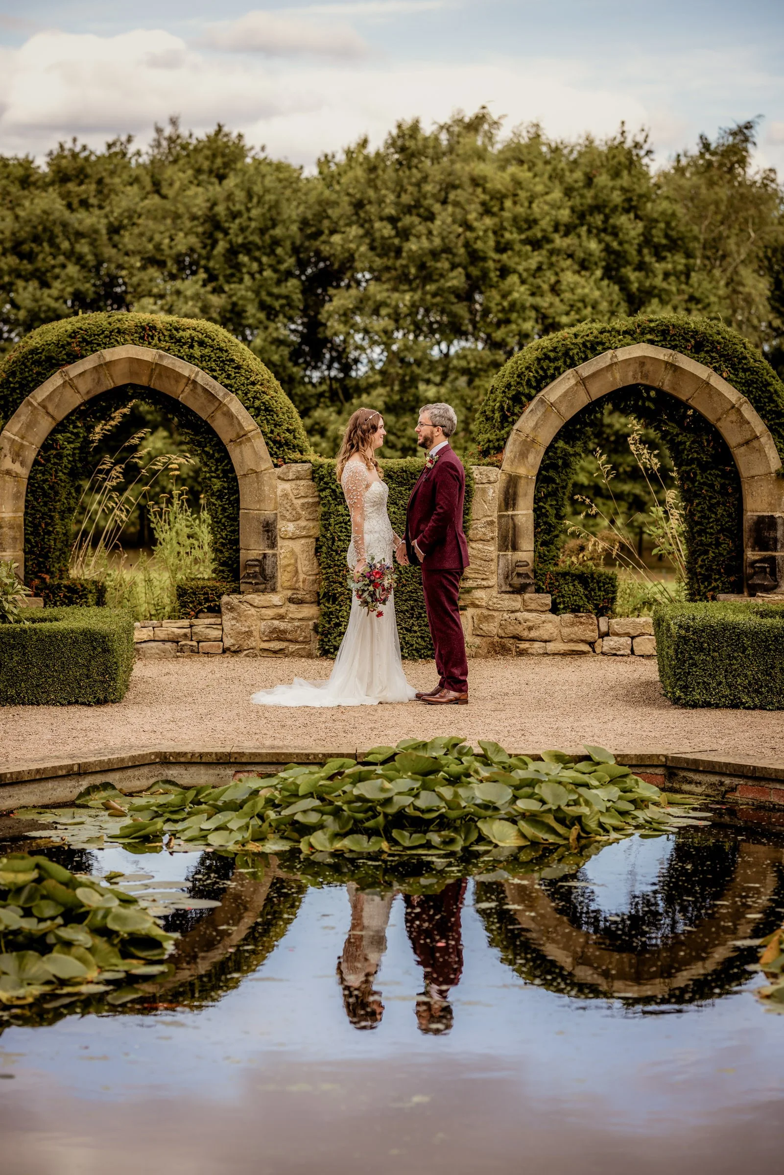 A bride and groom stand face-to-face in a garden with stone arches and lush greenery, reflected in a pond with lily pads.