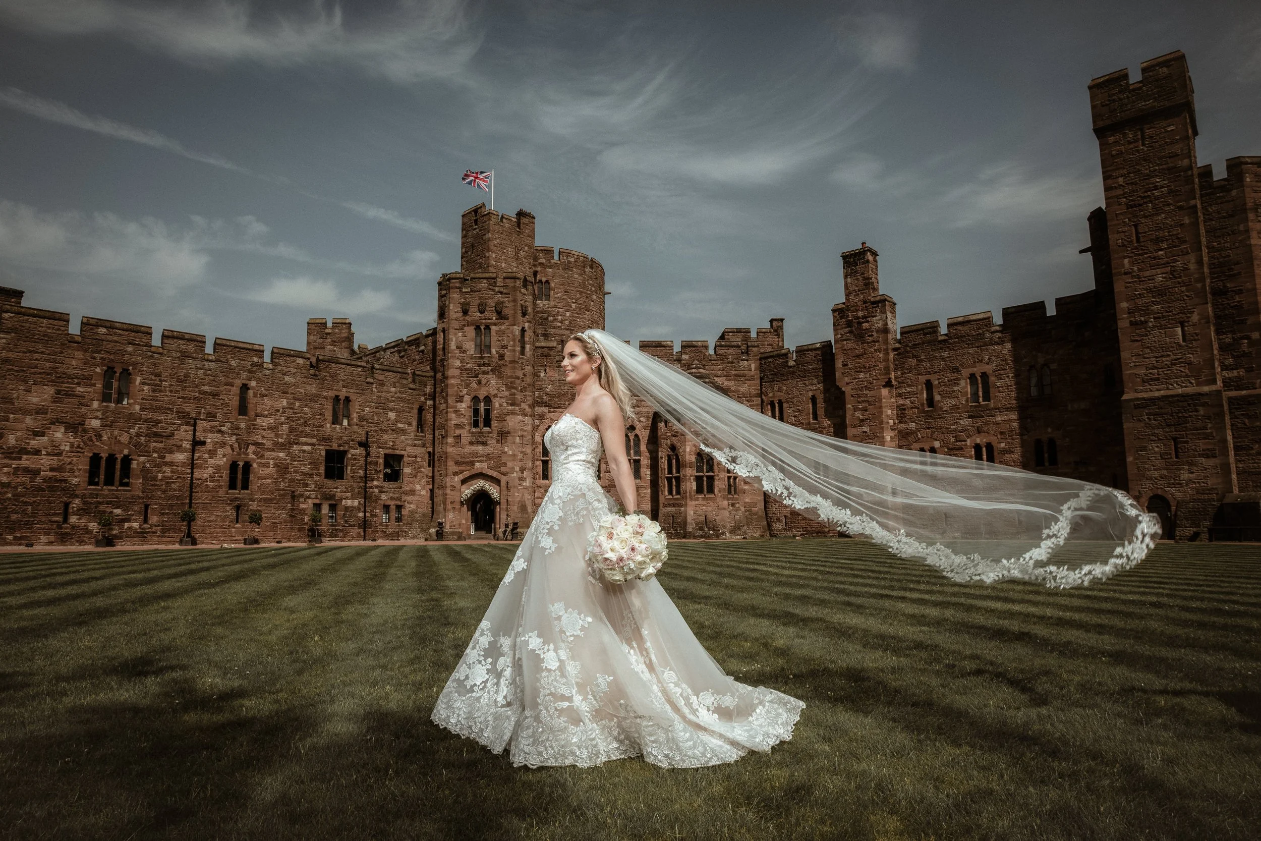 A bride in a wedding gown holding a bouquet of pink and white roses stands on a grassy area in front of a large historic castle with red brick walls and turrets, under a partly cloudy sky.