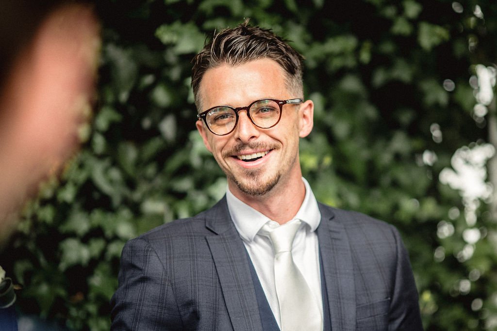 A man in a dark suit with a white shirt and tie, wearing glasses, smiling outdoors with greenery in the background.