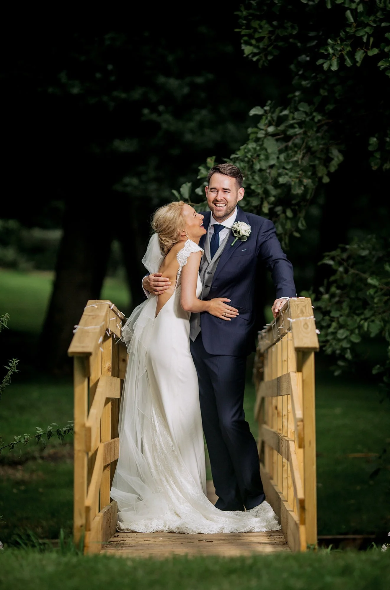 A bride and groom embrace and laugh on a wooden bridge outdoors, surrounded by greenery and trees.