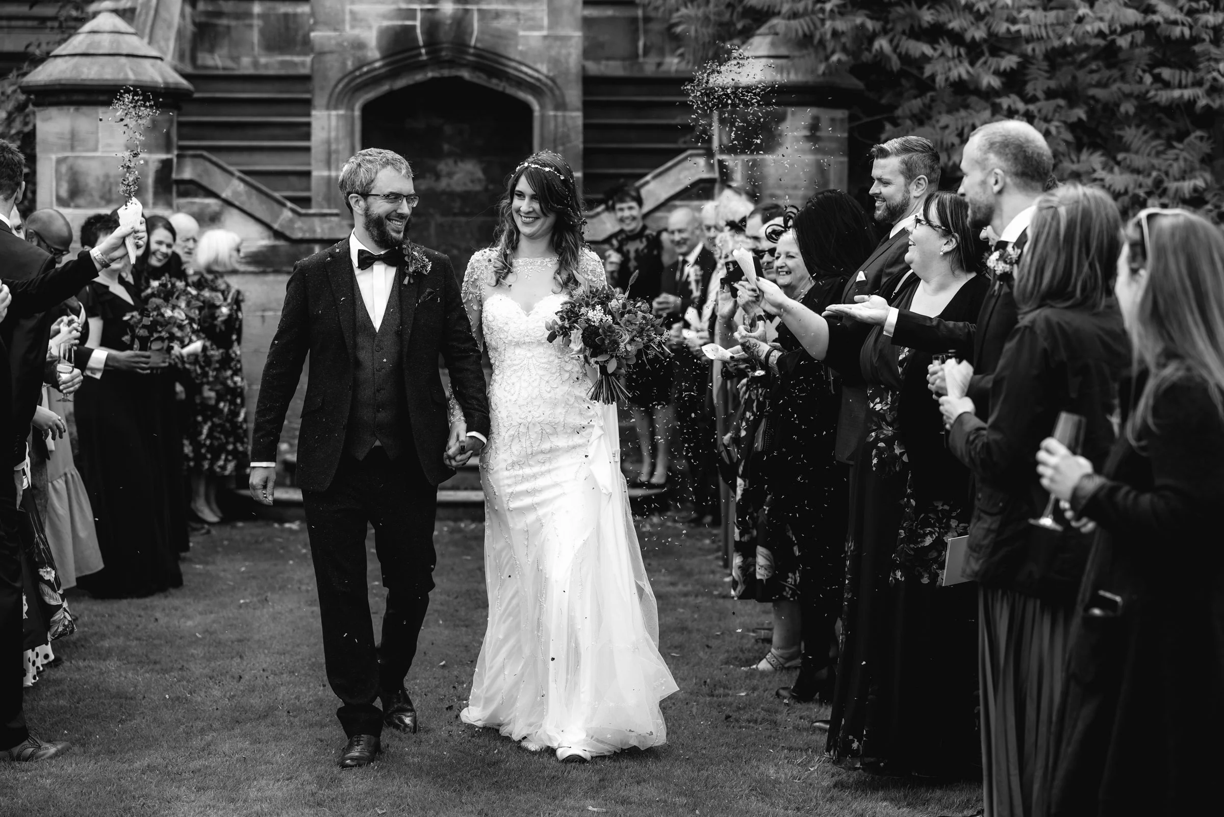 A black-and-white photo of a wedding celebration with a bride and groom walking hand in hand through a crowd of guests throwing confetti in front of an old stone building.