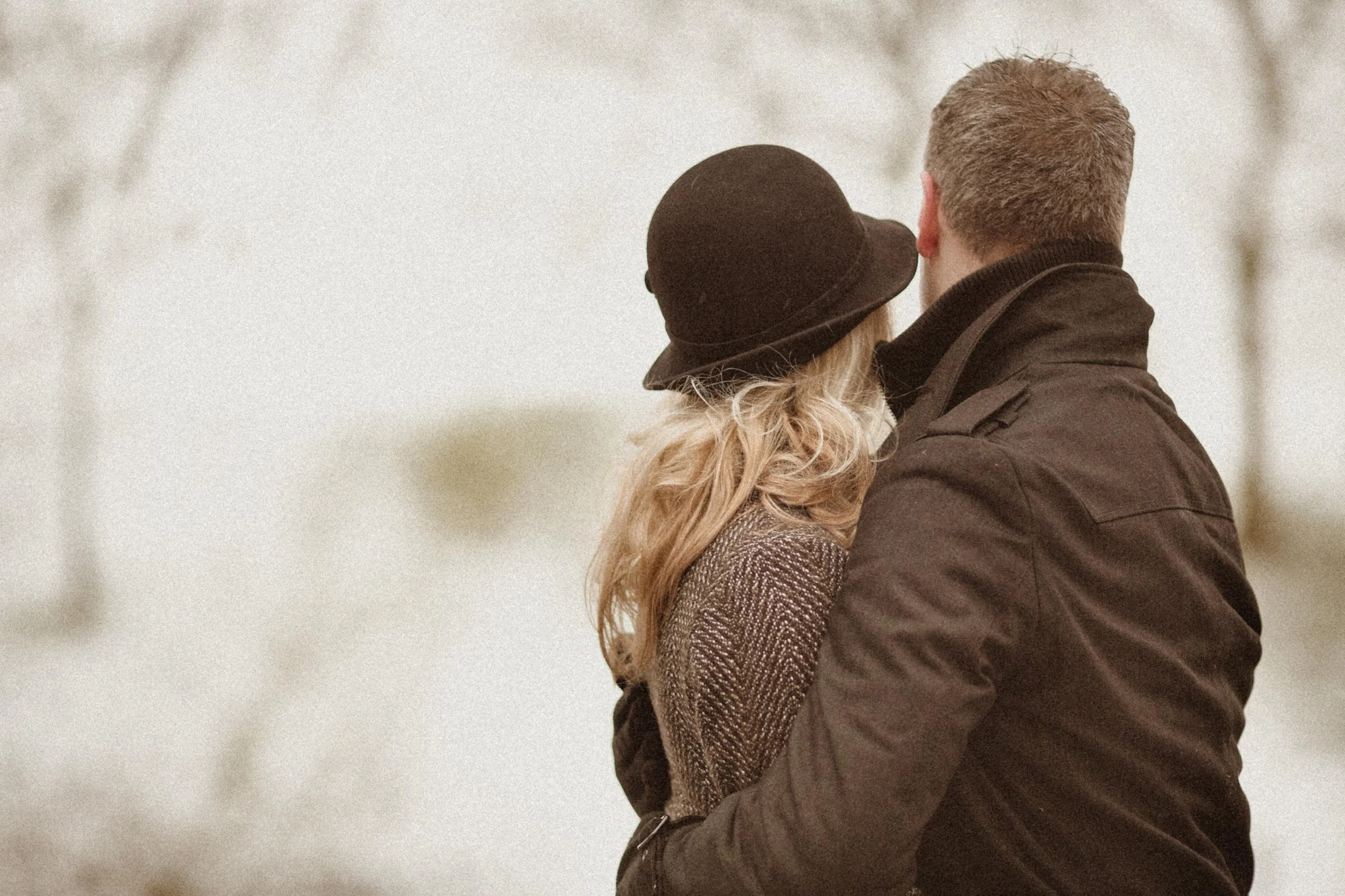 A couple embracing outdoors with blurred trees in the background, facing away from the camera.