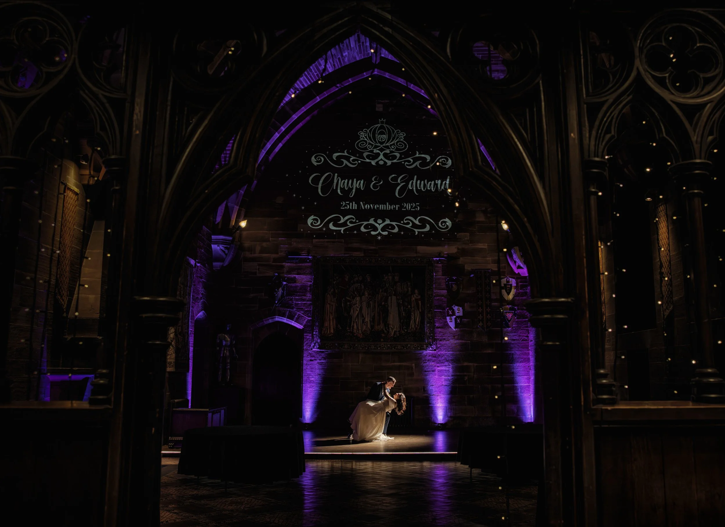 Couple dancing during a wedding reception inside a gothic-style hall with purple lighting, wood-paneled walls, and medieval decorations on the walls. A sign above reads 'Chaya & Edward, 25th November 2025'.