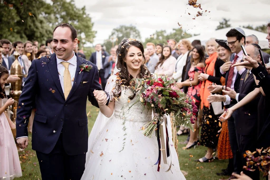 A bride and groom walking outdoors after their wedding, surrounded by smiling guests, with flower petals falling.