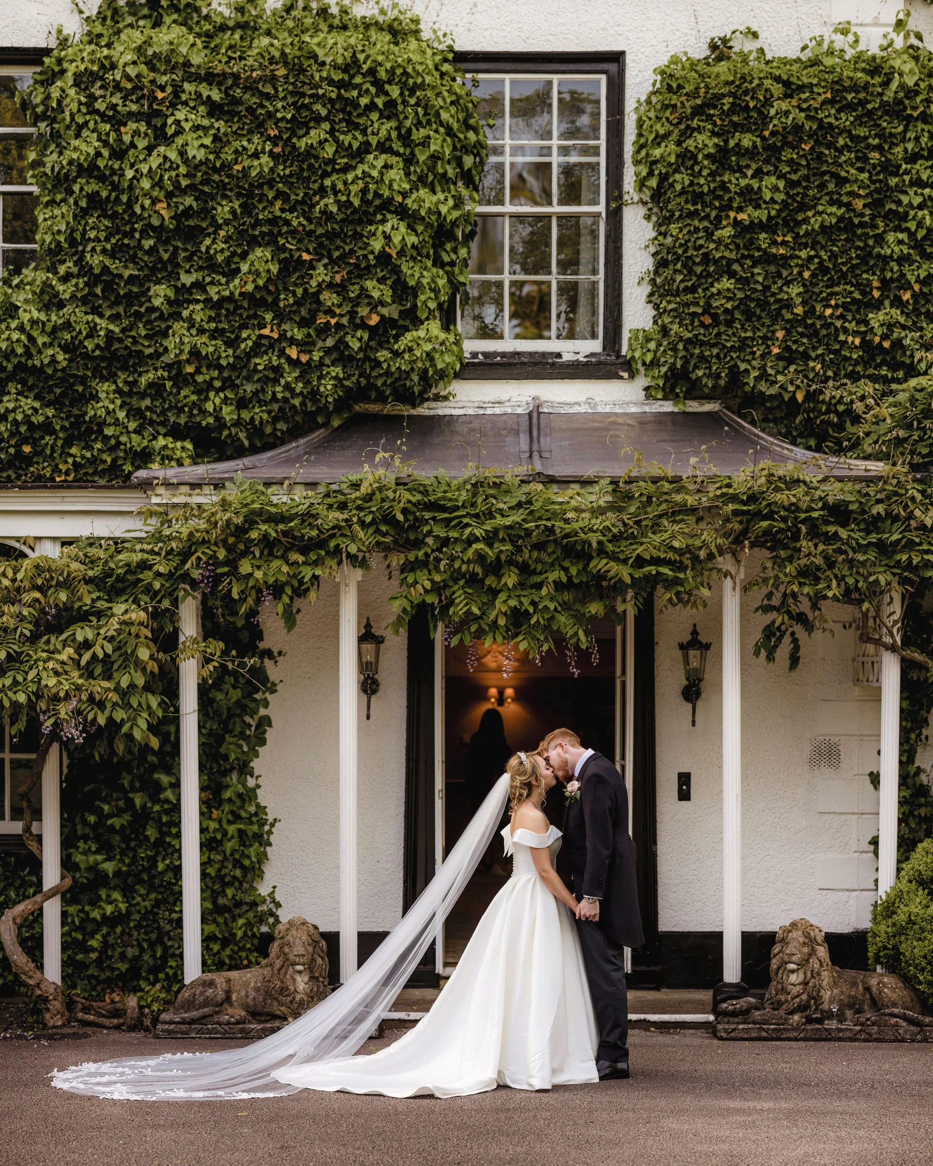 A bride and groom share a kiss in front of a white house with green ivy, lion statues, and a covered porch.
