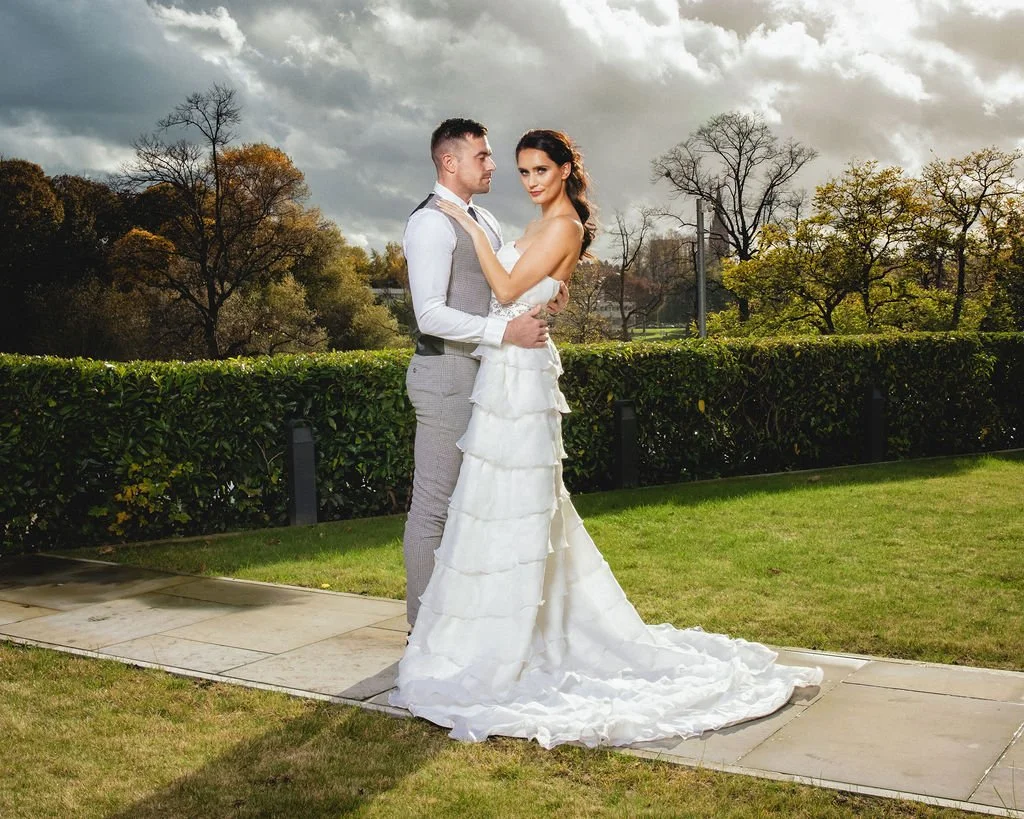 A bride and groom standing close together outdoors with trees and a cloudy sky in the background.