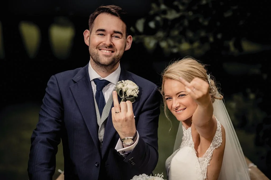 A bride and groom at their wedding, smiling and making playful gestures. The groom is in a navy suit holding a boutonniere, and the bride is in a lace wedding dress, making a rude hand gesture.
