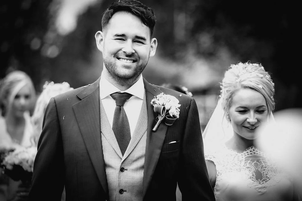 A black and white photo of a smiling groom in a suit with a boutonniere, standing close to a bride in a lace wedding dress and veil, at an outdoor wedding ceremony with guests in the background.
