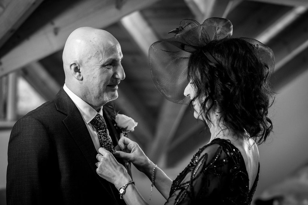 A woman wearing a large black hat and a black dress is pinning a boutonniere on a smiling bald man dressed in a suit. They are indoors with a wooden structure overhead.