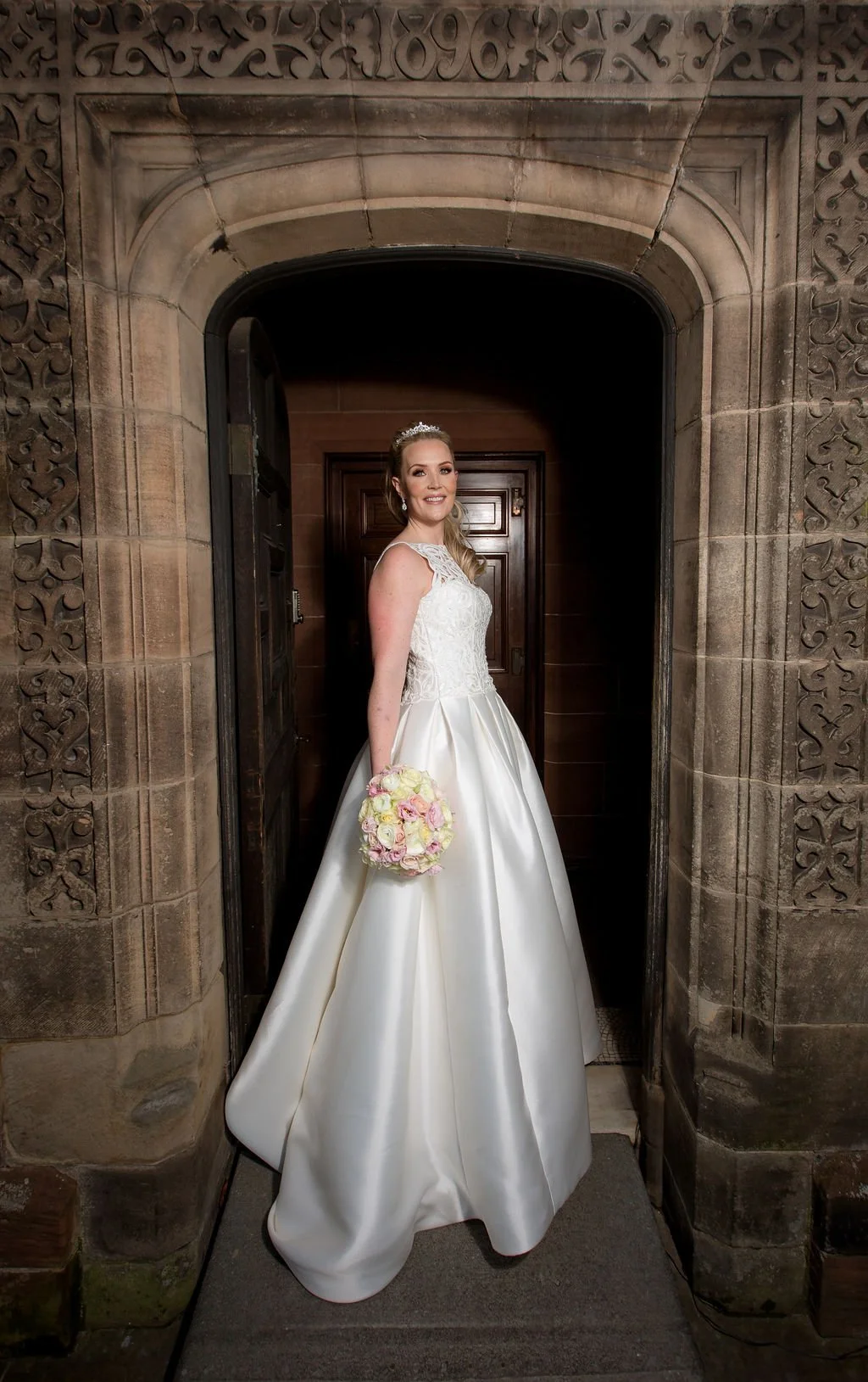 A bride in a white wedding gown holding a pink and white bouquet, standing in a stone doorway.
