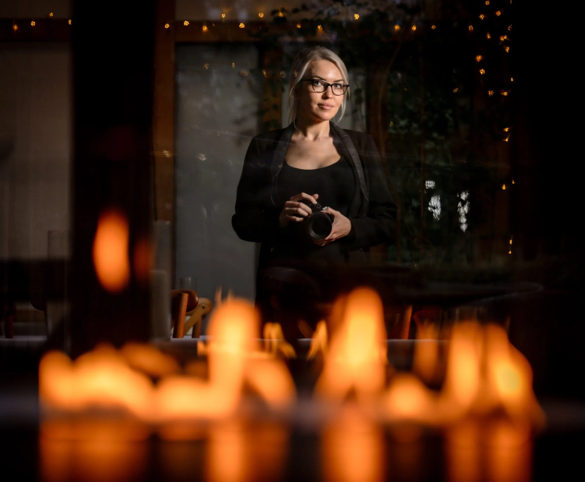 A woman with glasses and blonde hair takes a selfie or photographs through a glass window, with candles visibly burning in the foreground, creating a warm glow. She is wearing a black jacket and is holding a camera, with a background of outdoor string lights and trees.