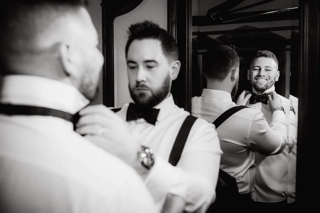 A man in formal attire adjusting his bow tie in front of a mirror, with the reflection showing him smiling as he prepares.