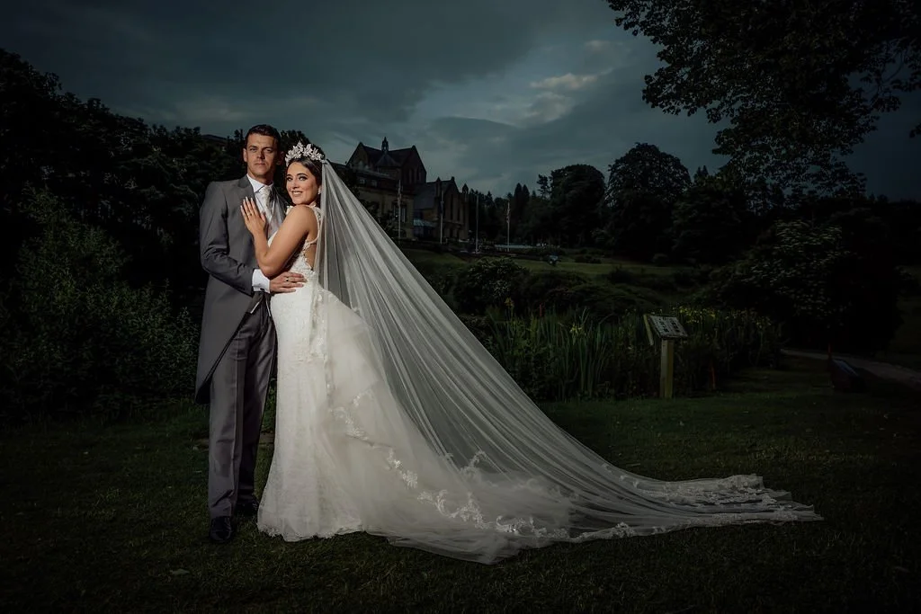 A bride and groom standing outdoors during dusk, with the bride in a white wedding dress and veil, and the groom in a gray suit. They are embracing, with a castle-like building and trees in the background.
