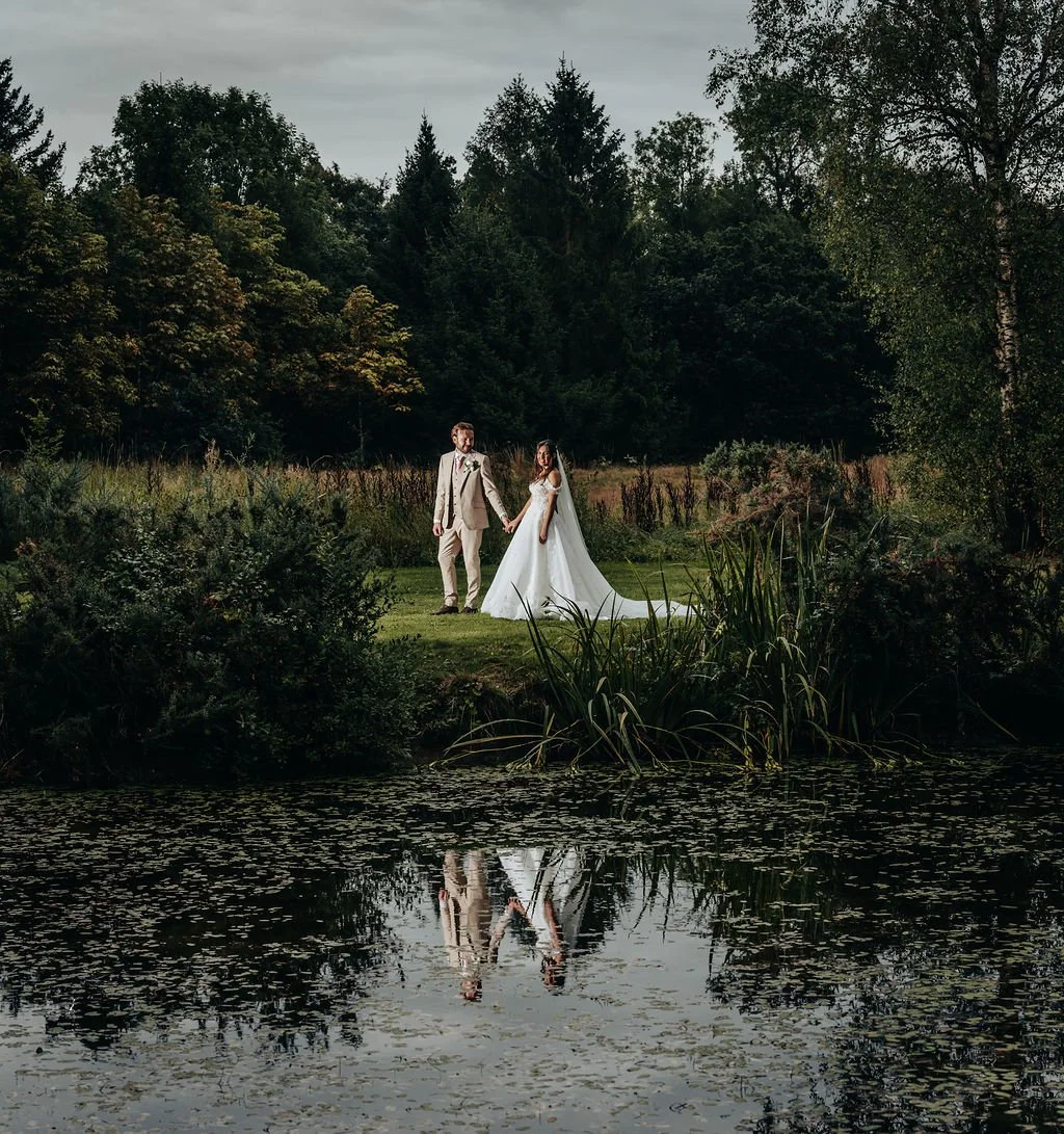 A bride and groom holding hands by a pond in a natural outdoor setting with trees and greenery.