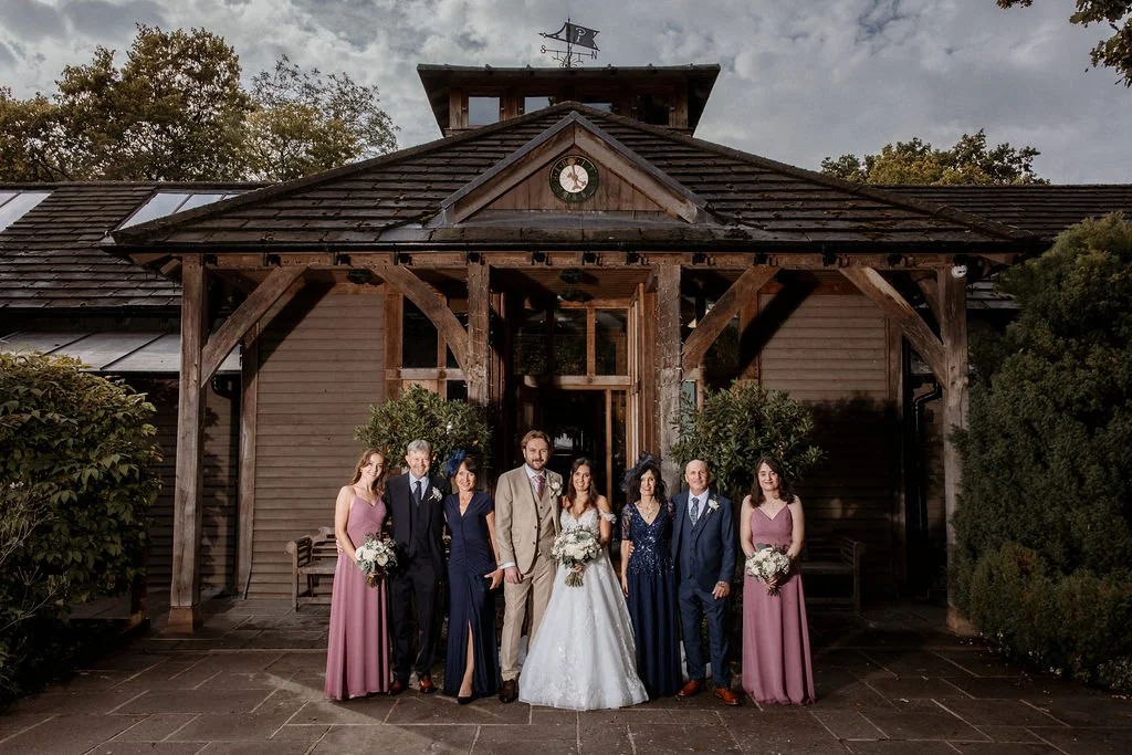 A wedding party standing in front of a wooden building with a clock, featuring the bride in a white gown, the groom in a tan suit, two bridesmaids in mauve dresses, and two groomsmen in suits, holding bouquets and smiling.