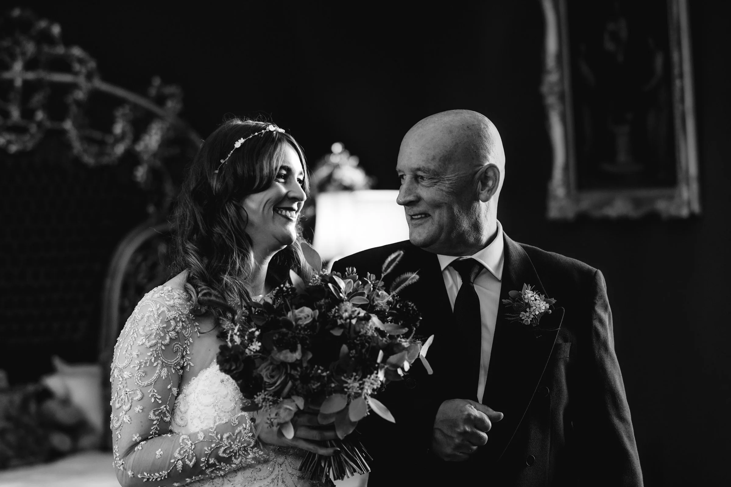 A bride in a lace dress holding a bouquet, smiling at a bald man in a suit, in a dimly lit room with a mirror and a lamp in the background.