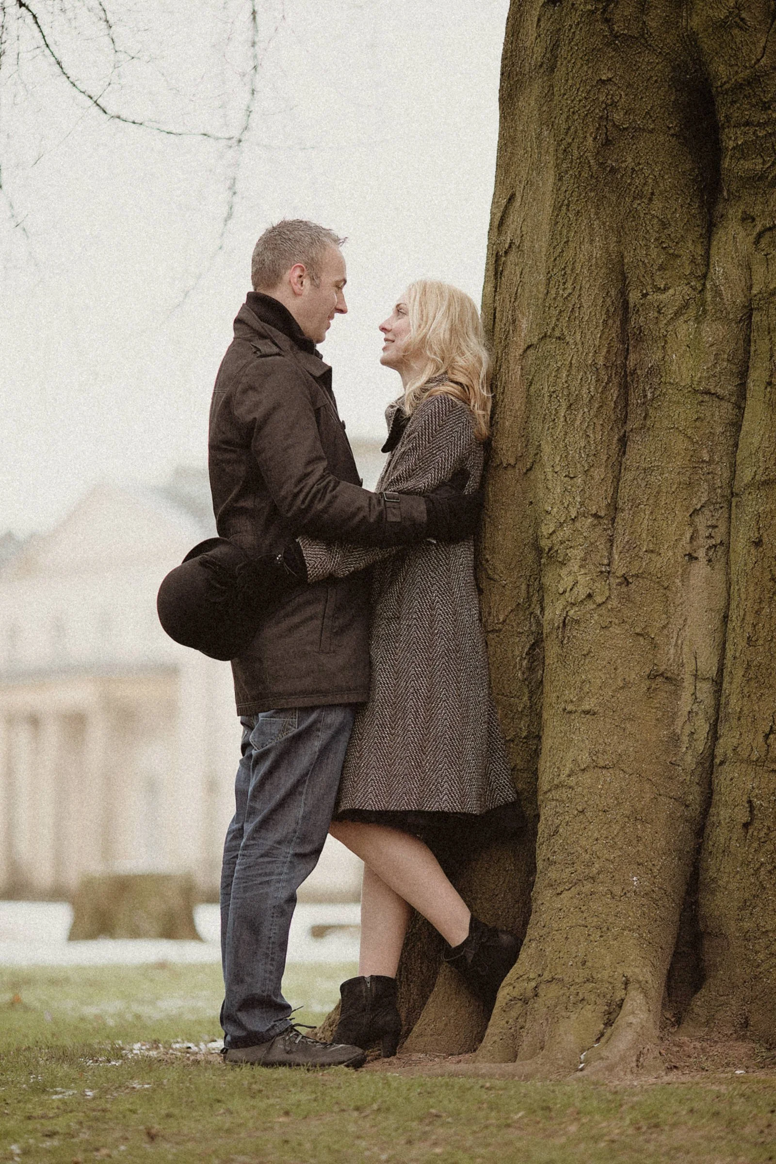 A couple standing close together outdoors, facing each other, with a large tree behind them. The woman leans against the tree, dressed in a coat and boots, while the man stands close, holding her arms.