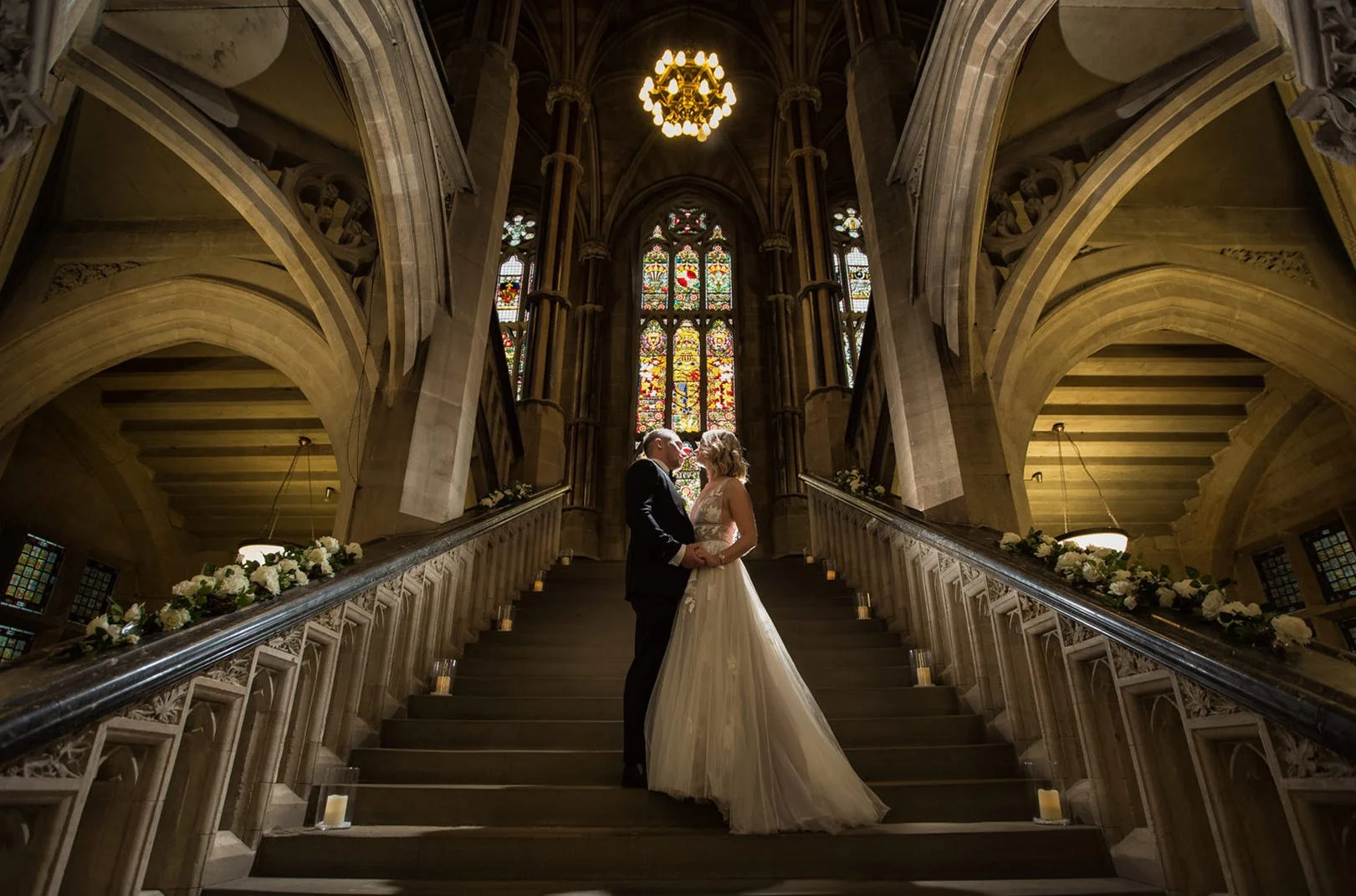 A bride and groom standing close and holding hands at the top of a staircase inside a grand church with stained glass windows, ornate stone arches, and decorated railing with flowers.