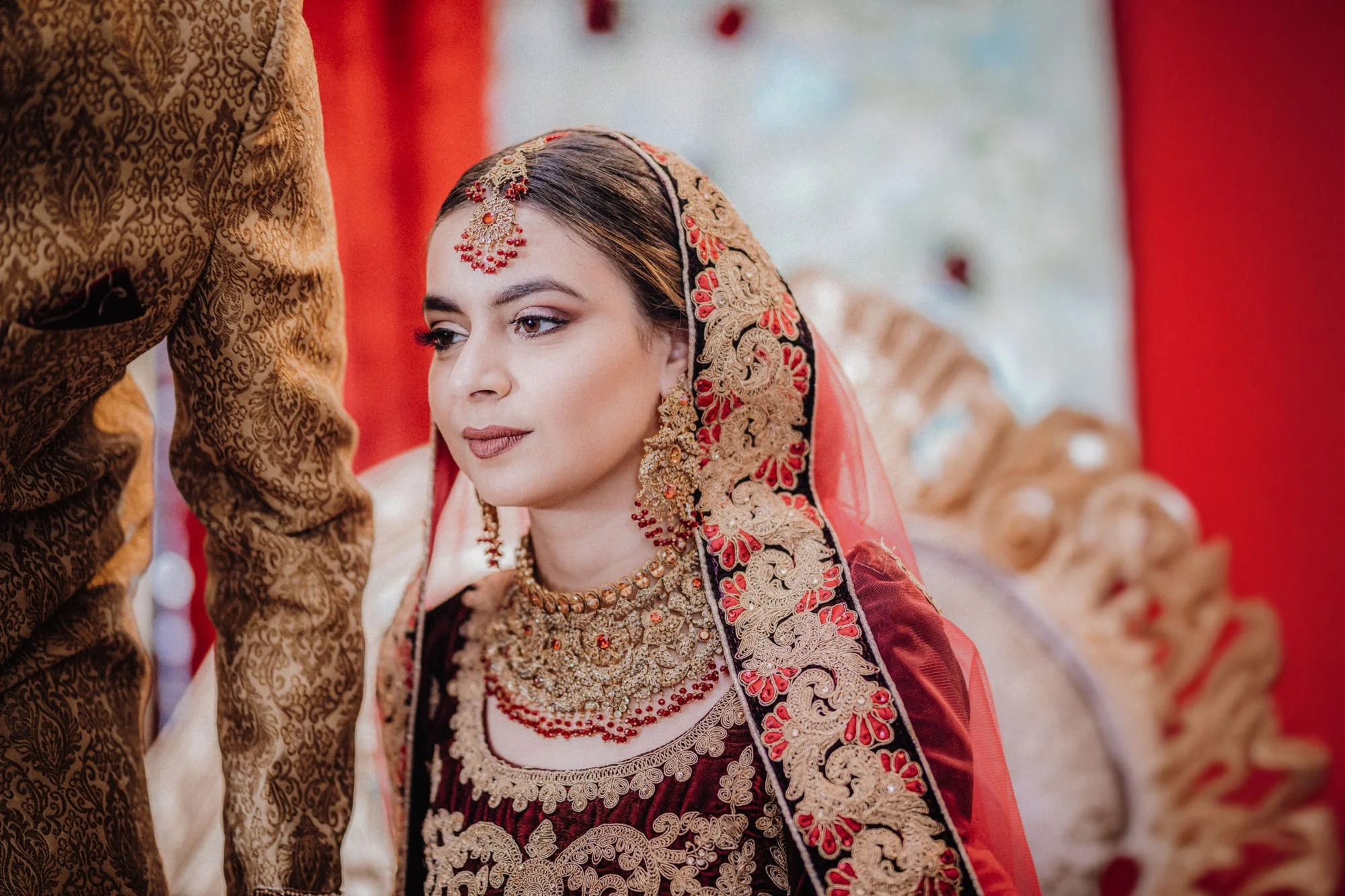 A bride dressed in traditional red and gold bridal attire with intricate embroidery, wearing elaborate jewelry including a necklace, earrings, and headpiece, during an Indian wedding ceremony.