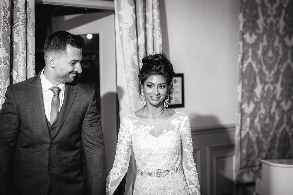 A bride in a lace wedding dress with earrings, holding hands with a groom in a suit, at a wedding ceremony.