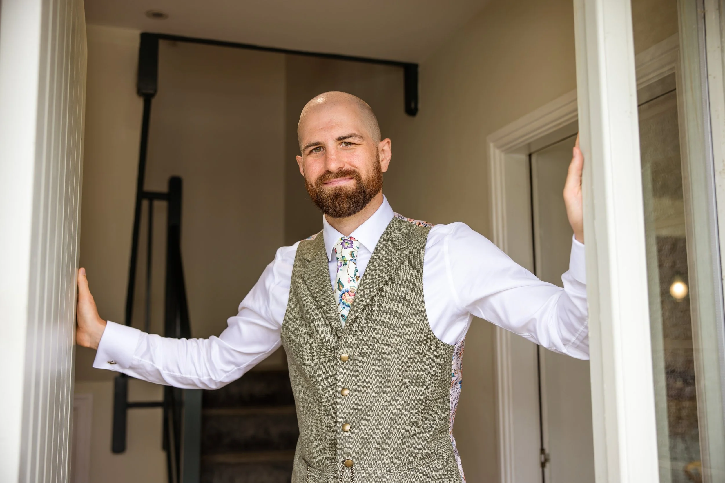 A man with a beard, dressed in a white shirt, vest, and colorful tie, standing at a doorway and smiling.