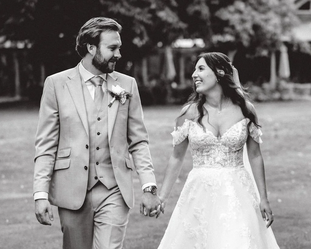 A black-and-white photo of a newlywed couple holding hands, smiling at each other outdoors on their wedding day, with trees and a building in the background.