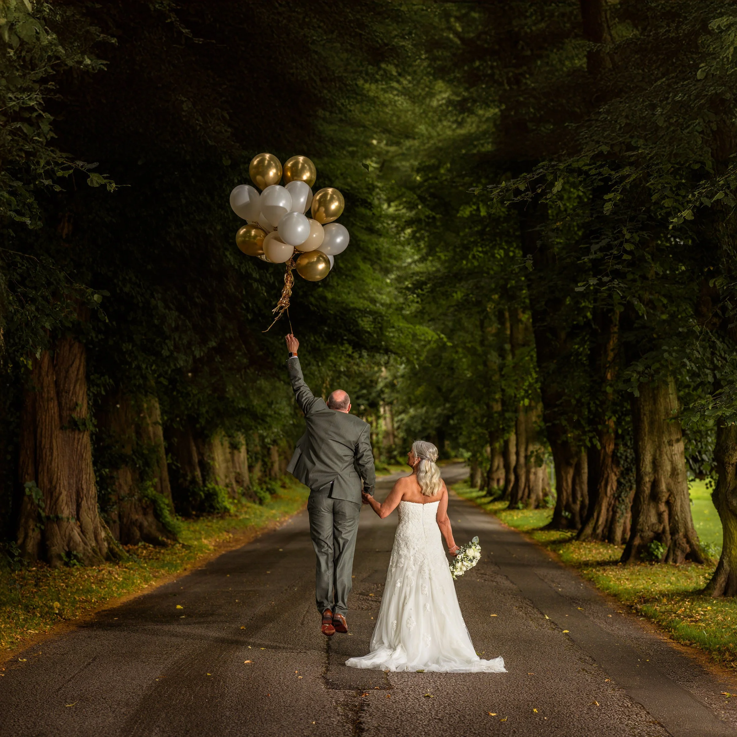 Groom holding balloons is lifted off the ground into the air whilst holding his brides hand on a drive way through the woods.