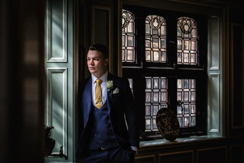 A young man in a formal black suit with a yellow tie, white shirt, and boutonniere stands by a window in an ornate interior, looking out thoughtfully.