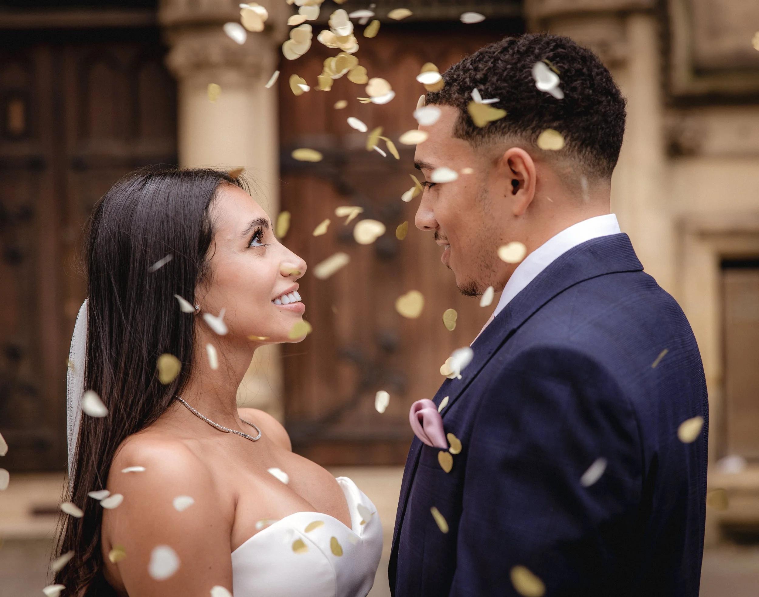 A bride and groom gazing into each other's eyes surrounded by falling gold and white confetti during their wedding celebration.