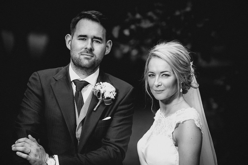 Black and white photo of a bride and groom on their wedding day, both looking at the camera. The groom wears a suit with a boutonniere, and the bride wears a lace wedding dress with a veil.