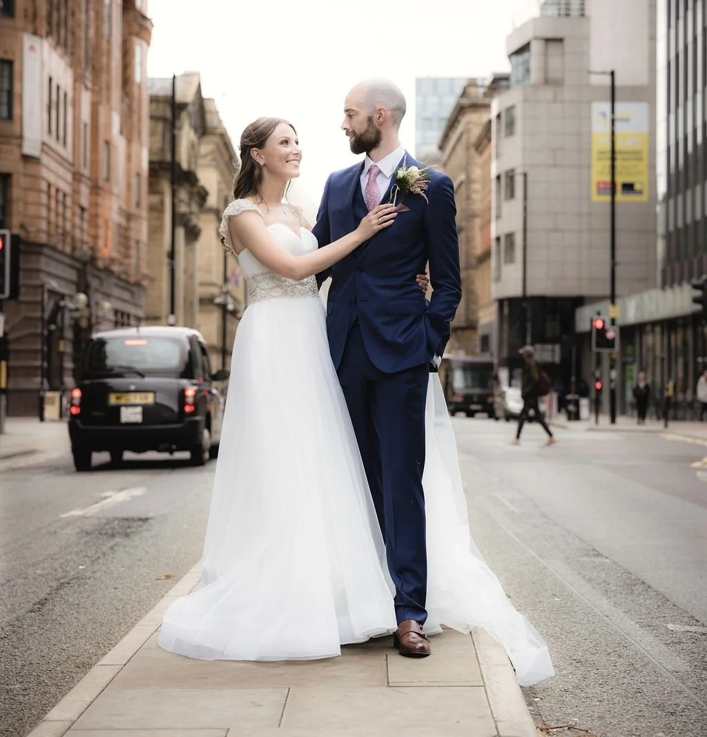 A bride and groom dressed in wedding attire on a city street, gazing at each other, with buildings and cars in the background.