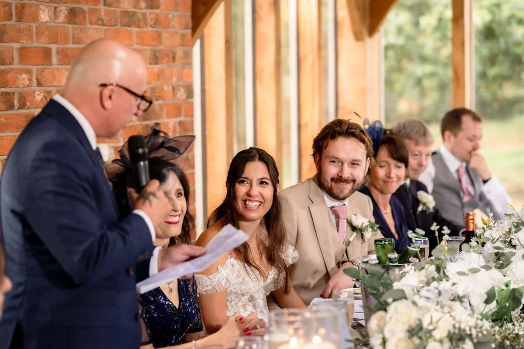 People sitting at a long table during a wedding reception, listening to a man in a suit giving a speech, with a brick wall and window in the background.