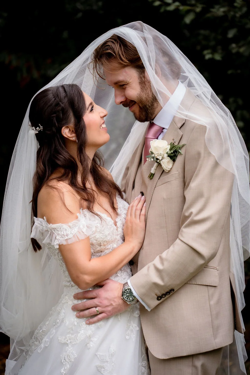 A bride and groom smile while looking into each other's eyes, close together, during a wedding photo. The bride has long dark hair with a decorative hairpiece and is wearing an off-the-shoulder lace wedding dress. The groom has light brown hair, a be