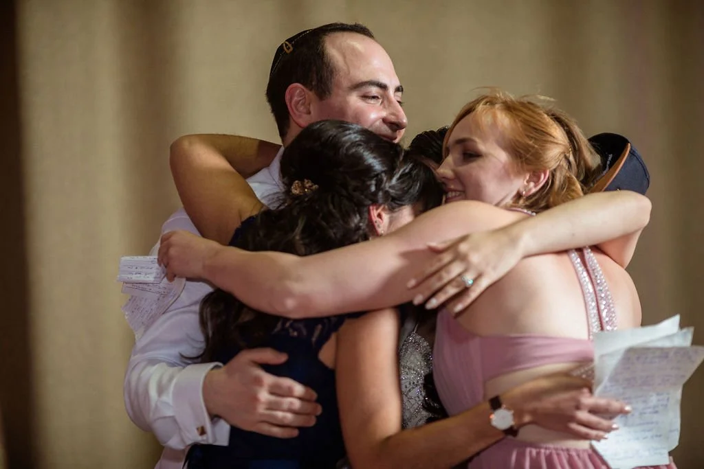 Four people hugging each other tightly in a joyful group hug, celebrating a special moment, possibly at a wedding or family gathering.