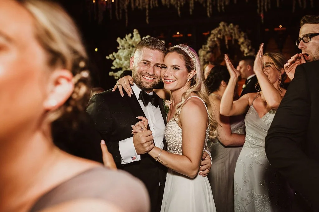 A bride and groom dancing closely at their wedding reception, smiling and surrounded by guests.