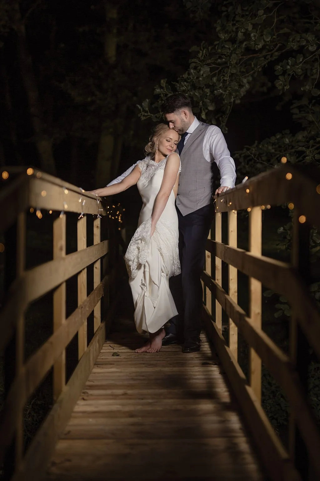 A bride and groom standing close together on a small wooden bridge at night, with string lights decorating the bridge railings, surrounded by dark trees.