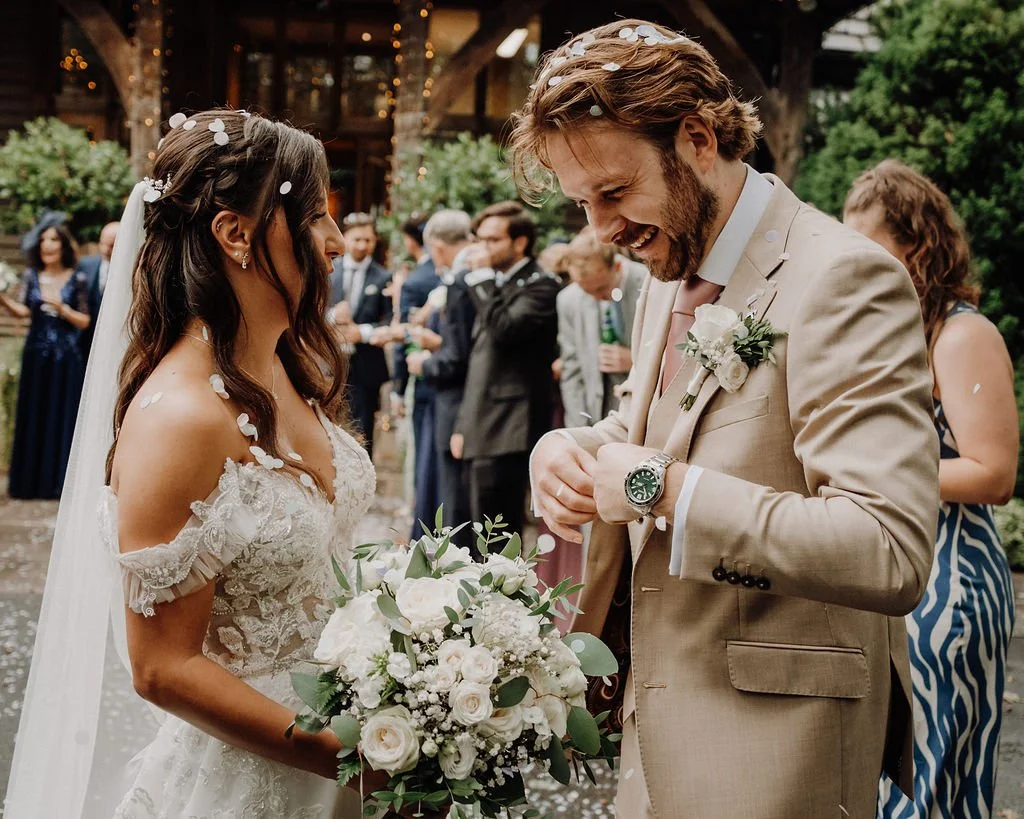 A bride and groom are exchanging vows at their outdoor wedding ceremony, surrounded by guests. The bride is holding a bouquet of white flowers, and the groom is smiling as he checks his watch.