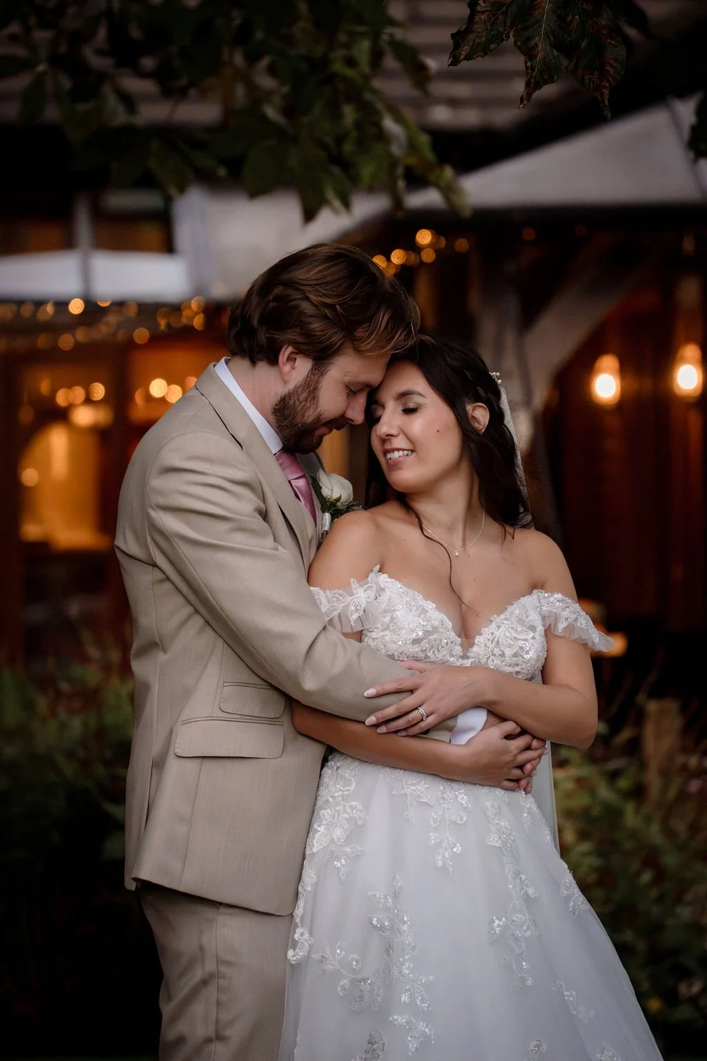 A newlywed couple sharing a romantic moment outdoors during their wedding reception, with warm lighting and string lights in the background.